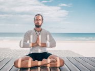 Adult bearded man meditating while sitting in lotus pose on wooden pier by seashore with legs crossed and looking at camera — Stock Photo