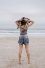 Back view of attractive woman wearing top and shorts standing on sandy seashore with black hat — Stock Photo