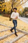 Гламурный красивый мужчина в джинсах и в белом свитере ходит по желтым осенним листьям в городе — стоковое фото