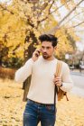 Hombre joven guapo con peinado de moda caminando sobre hojas de otoño y hablando en el teléfono inteligente en el día - foto de stock
