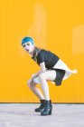 Sensuale giovane donna in abito futuristico alla moda guardando la fotocamera mentre in piedi sulle ginocchia sul marciapiede contro parete gialla brillante — Foto stock