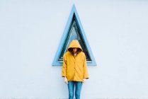 Young woman in yellow warm coat smiling and looking down while standing against triangle window and gray wall of building — Stock Photo