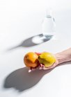 Gros plan de la main femelle tenant orange mûr et citron près de pot clair d'eau sur fond blanc — Photo de stock