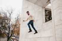 Trendy young man in jeans and white sweater running in air at white building — Stock Photo