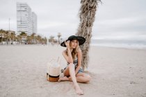 Блондинка в чорному капелюсі сидить на піску з літньою сумкою і дивиться на камеру — стокове фото