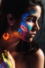 Seitenansicht der schönen jungen Frau, die mit Leuchtfarbe im Gesicht, Hals und Hand bedeckt ist und wegschaut — Stockfoto