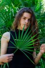 Portrait of young brunette woman in tropical bushes holding palm tree leaf — Stock Photo