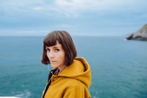 Жінка в жовтій теплій куртці, дивлячись на камеру, стоячи на тлі розриву моря і похмурого неба в природі — стокове фото