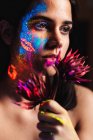 Retrato de bela jovem coberta com tinta luminosa no rosto segurando uma flor e olhando para longe — Fotografia de Stock