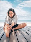 Erwachsener bärtiger Mann streckt sich, während er auf einem hölzernen Steg am Meer sitzt und wegschaut — Stockfoto