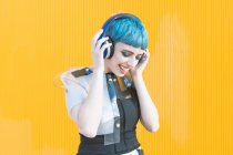 Joyeux jeune femme en robe alternative à la mode souriant et écoutant de la musique dans les écouteurs contre le mur jaune — Photo de stock