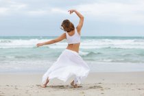 Back view of unrecognizable female in white outfit dancing on sand near waving sea — Stock Photo
