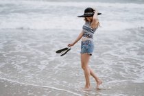 Femme attrayante en chapeau noir tenant des chaussures tout en profitant d'une vue pittoresque sur l'océan regardant vers le bas — Photo de stock