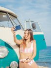 Mulher bronzeada encantadora atraente levando selfie e sorrindo perto de carro na praia de areia em dia brilhante — Fotografia de Stock