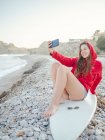 Junge lächelnde Frau mit langen Haaren in rotem Kapuzenpulli macht Selfie mit dem Smartphone auf dem Surfbrett am steinigen Meer liegend — Stockfoto