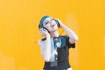Allegro giovane donna in abito alternativo alla moda sorridente e ascoltare musica in cuffia contro il muro giallo — Foto stock