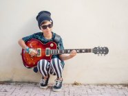 Cheeky ragazzo eccitato attivo in vestiti colorati suonare la chitarra, appoggiato sullo sfondo della parete bianca — Foto stock