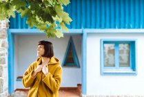 Jeune femme en manteau jaune chaud souriant et regardant loin tout en se tenant contre le bâtiment mural gris — Photo de stock