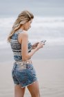 Вид ззаду молода блондинка весела жінка стоїть і використовує смартфон на морському фоні — стокове фото