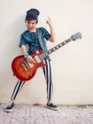 Нахабний активний хлопчик в барвистому одязі грає на гітарі, показуючи два пальці вгору на тлі білої стіни — стокове фото
