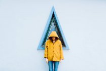 Junge Frau in gelbem warmen Mantel lächelt und schaut nach unten, während sie vor einem Dreiecksfenster und einer grauen Hauswand steht — Stockfoto