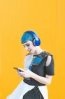 Fröhliche, ungezwungene Frau mit Kopfhörern, die auf ihrem Smartphone surft und Musik hört, während sie vor einer leuchtend gelben Wand steht — Stockfoto