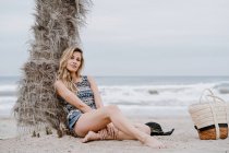 Portrait of young beautiful blonde woman sitting down on beach and looking at camera — Stock Photo