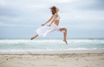 Atractiva hembra en traje blanco bailando en la arena cerca del mar ondulante - foto de stock