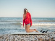 Vue latérale d'une jolie femme aux cheveux longs et souriants en sweat à capuche rouge appréciant de s'asseoir sur une planche de surf sur une plage pierreuse — Photo de stock