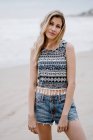 Cheerful blonde woman in colorful top and jean shorts posing while relaxing on seashore — Stock Photo