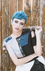 Giovane donna con i capelli corti blu indossando abito futuristico e guardando la fotocamera mentre in piedi vicino alla recinzione in legno shabby — Foto stock