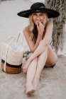 Блондинка в черной шляпе сидит на песке с летней сумкой и смотрит в камеру — стоковое фото