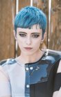 Junge Frau mit kurzen blauen Haaren trägt futuristisches Kleid und blickt in die Kamera, während sie am schäbigen Bauzaun steht — Stockfoto