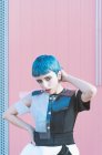 Молодая женщина в модном альтернативном платье трогает короткие голубые волосы и смотрит в камеру, стоя напротив розовой стены — стоковое фото
