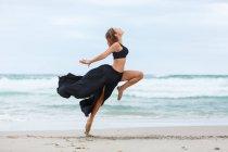 Attractive female in black outfit dancing on sand near waving sea — Stock Photo
