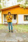 Young woman in casual outfit smiling and browsing smartphone while standing on tiled path outside lovely cottage on autumn day in countryside — Stock Photo