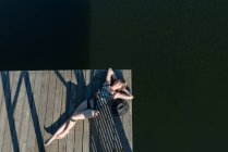 Draufsicht der schönen Frau in schwarzem Badeanzug und Hut liegend auf Holzpier des Sees bei klarem blauem Himmel und Waldhintergrund — Stockfoto