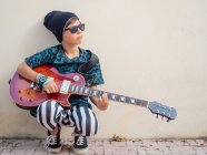 Cheeky actif excité garçon en vêtements colorés jouer de la guitare, appuyé sur le fond de mur blanc — Photo de stock