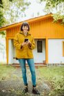 Mujer joven en traje casual sonriendo y navegando teléfono inteligente, mientras que de pie en el camino de baldosas fuera encantadora casa de campo en el día de otoño en el campo - foto de stock