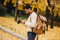 Beau jeune photographe debout dans le parc d'automne et tenant la caméra — Photo de stock