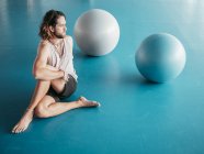 Desde arriba de hombre barbudo en ropa deportiva estiramiento con las piernas cruzadas en el suelo azul con bolas de gimnasia en el estudio - foto de stock