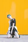 Junge Frau im trendigen futuristischen Kleid blickt in die Kamera, während sie auf Knien auf dem Bürgersteig vor einer knallgelben Wand sitzt — Stockfoto