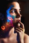 Side view of beautiful young woman covered with luminous paint on face touching lips with finger — Stock Photo
