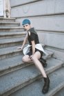 Mujer joven con el pelo azul corto con vestido informal de moda y posando en las escaleras de la calle - foto de stock