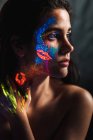 Porträt einer schönen jungen Frau, die mit Leuchtfarbe im Gesicht, am Hals und an der Hand bedeckt ist und wegschaut — Stockfoto