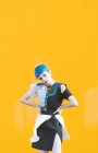 Giovane donna in abito futuristico alla moda guardando la fotocamera mentre in piedi sulle ginocchia sul marciapiede contro la parete gialla brillante — Foto stock