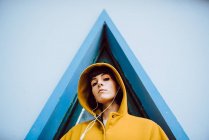 Junge Frau in gelbem warmen Mantel, die Musik hört und in die Kamera schaut, während sie vor einem Dreiecksfenster und einer grauen Hauswand steht — Stockfoto