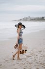Atractiva mujer en sombrero negro sosteniendo bolsa de playa y zapatos mientras disfruta de una pintoresca vista del océano - foto de stock