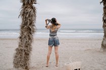 Back view of woman wearing top and shorts standing on sandy seashore with black hat — Stock Photo