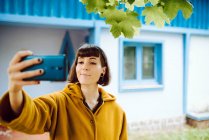 Mujer joven en chaqueta cálida amarilla sonriendo y utilizando el teléfono inteligente para tomar selfie en el fondo borroso de la casa de campo - foto de stock